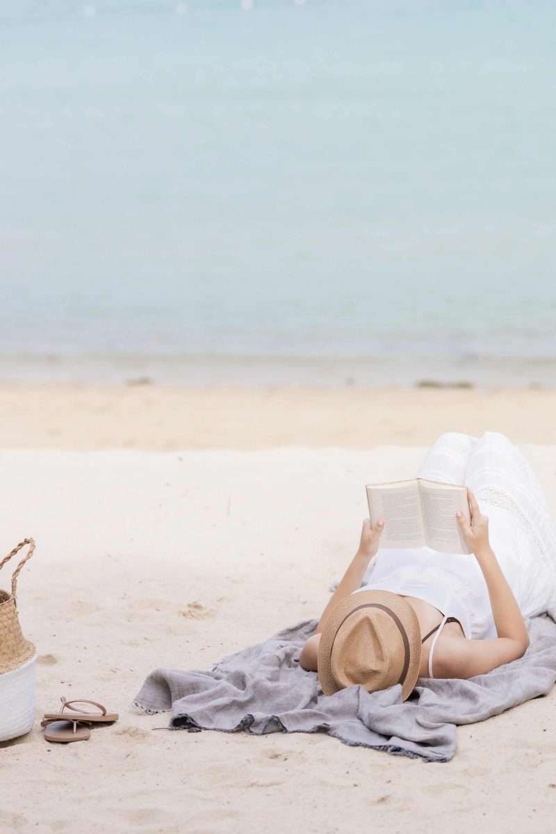 Using the Envelope System: A Beginner’s Budgeting Guide Woman in a white dress laying on a gray beach towel and a beach reading a book with flip-flops and a purse. 