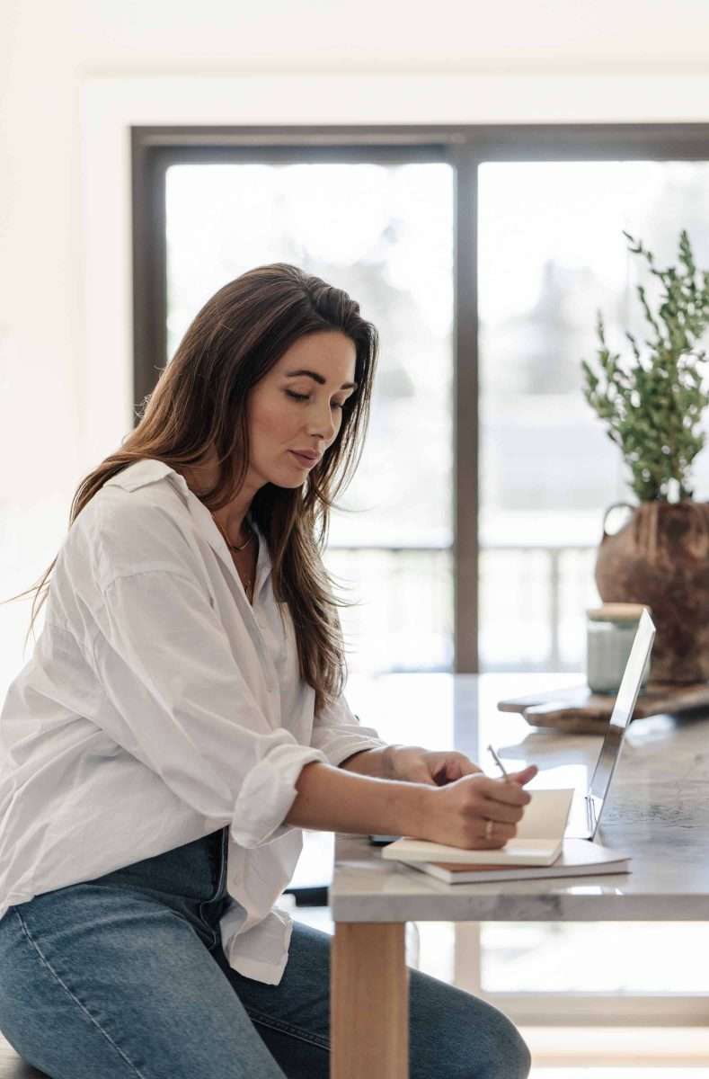Using the Envelope System: A Beginner’s Budgeting Guide Woman in a white shirt and blue jeans and a kitchen counter writing in her journal with a laptop.