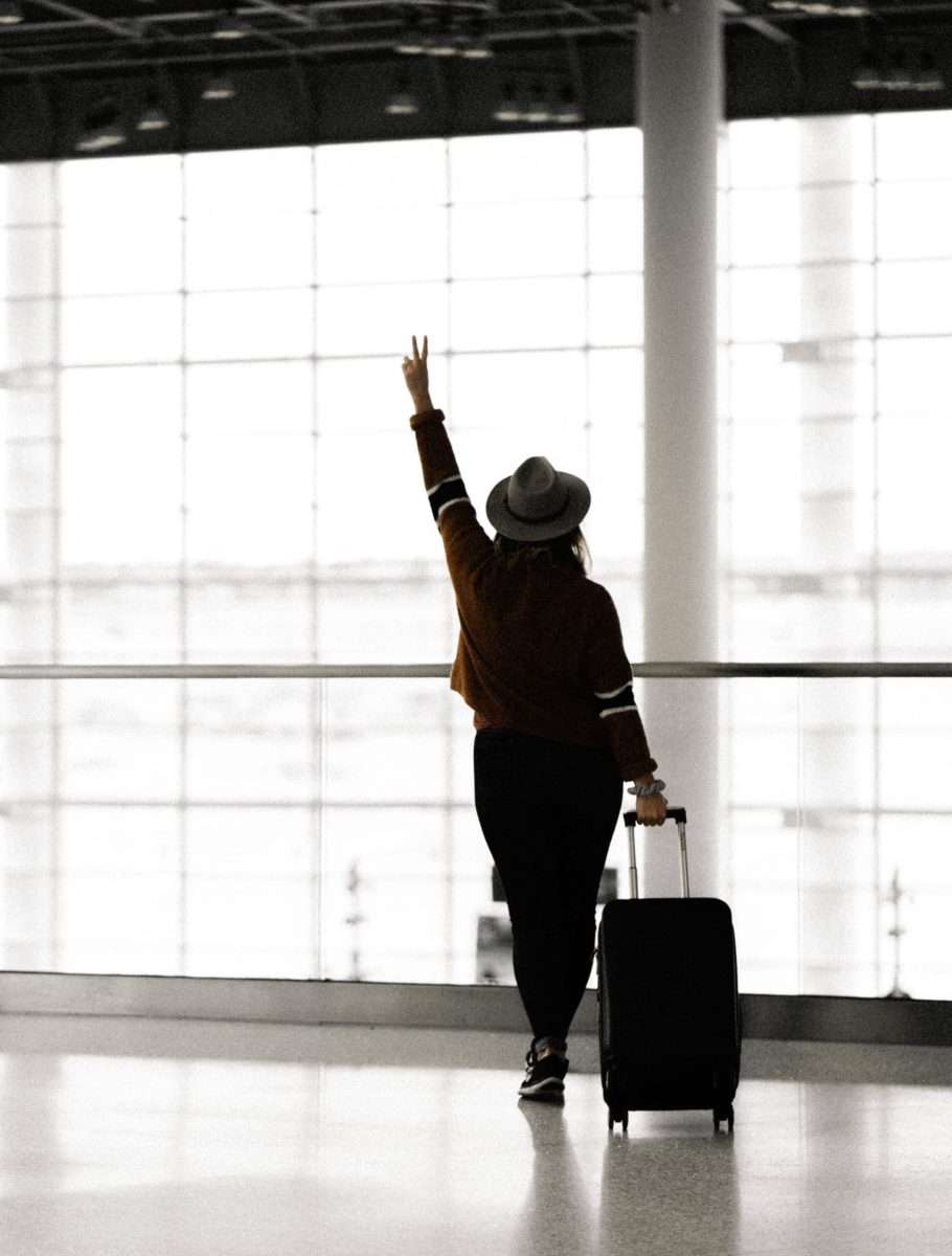 Using the Envelope System: A Beginner’s Budgeting Guide woman with a hat and a suitcase with back facing the camera left arm up with the victory sign in an airport leaving for vacation. 