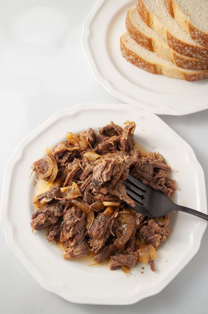 top down view of teriyaki flank steak slow cooker white plate with a pile of shredded flank steak with bits of onion on a black serving fork and on another white plate 4 slices of bread crockpot recipe
