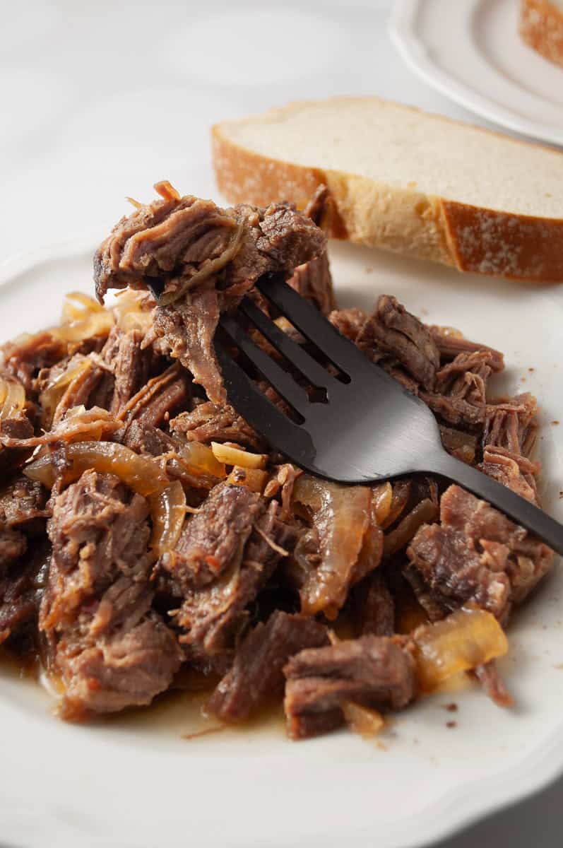 teriyaki flank steak slow cooker white plate with a pile of shredded flank steak with bits of onion on a black serving fork with a slice of bread crockpot recipe