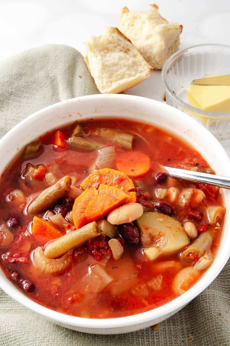 Healthy CrockPot Vegetable Minestrone Soup in a white bowl on a green towel and a spoon at the top a small bowl with butter and a roll of a towel.