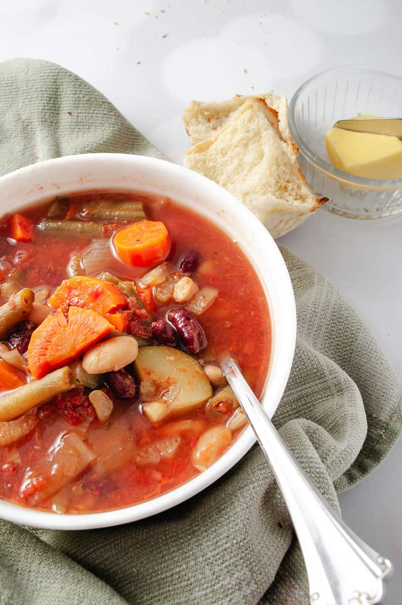 Healthy CrockPot Vegetable Minestrone Soup in a white bowl on a green towel and a spoon at the top a small bowl with butter and a roll of a towel.