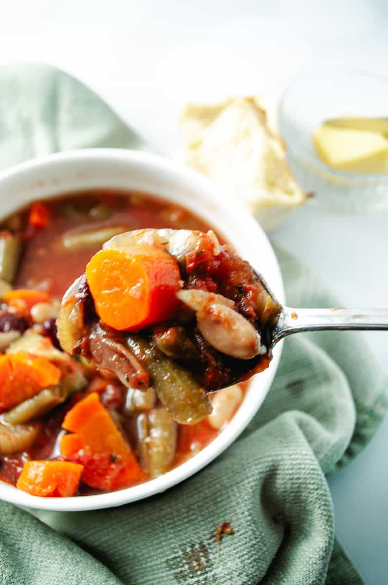 Healthy CrockPot Vegetable Minestrone Soup in a white bowl on a green towel and a spoon at the top a small bowl with butter and a roll of a towel close up of the soup in the spoon.