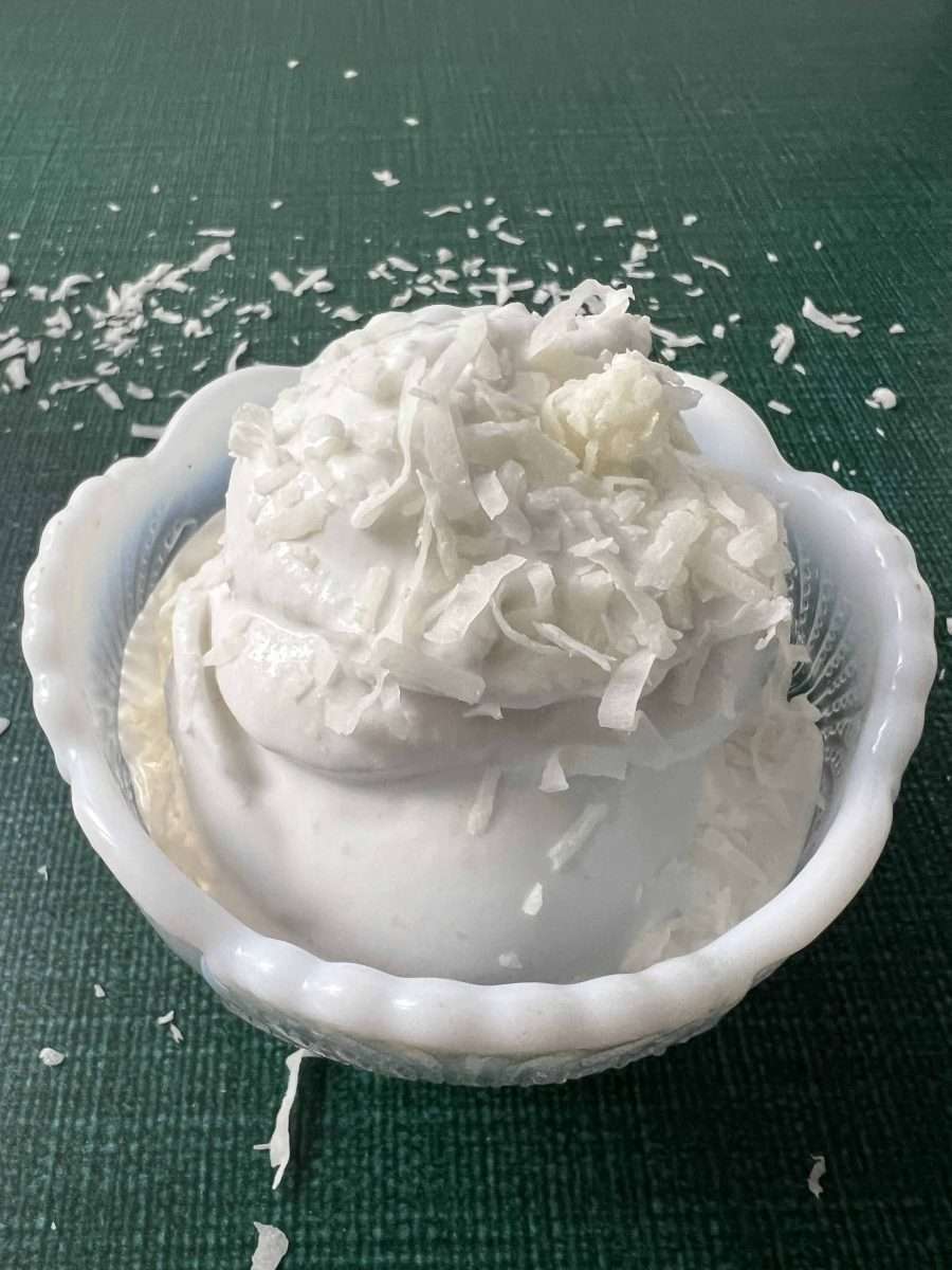 Ninja Creami Lite Coconut Ice Cream white bowl of ice cream in a green counter with flakes of coconut on the ice cream and the counter.