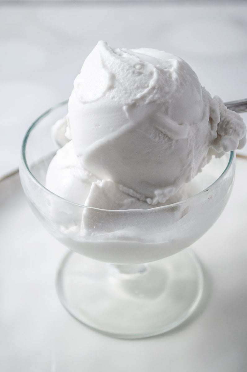Dairy-Free Vanilla Coconut Ice Cream Ninja Creami clean ice cream parfait cup with 2 scoops of creamy coconut ice cream with a spoon and sitting on a white plate with a gold trim.