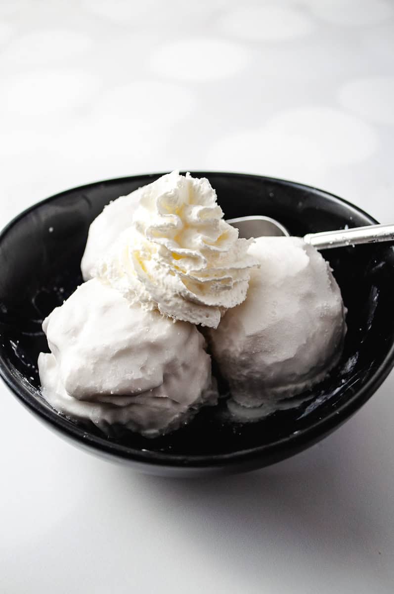 Dairy-Free Vanilla Coconut Ice Cream Ninja Creami clean ice cream parfait cup with 2 scoops of creamy coconut ice cream with a spoon and sitting on a white plate with a gold trim.