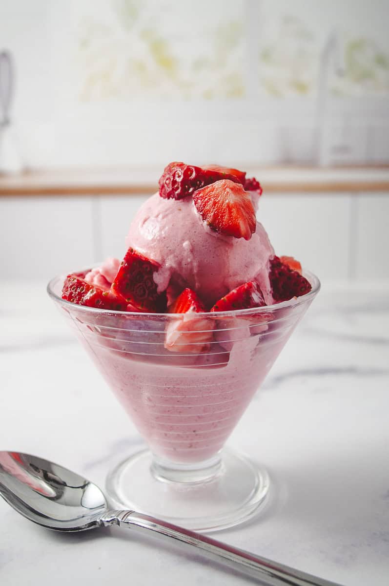 Ninja Ceami Protein Strawberry Ice Cream with Fairlife Milk in a decorative serving cup with cut up fresh strawberries on top, on a white counter with a spoon in front up close of the ice cream.