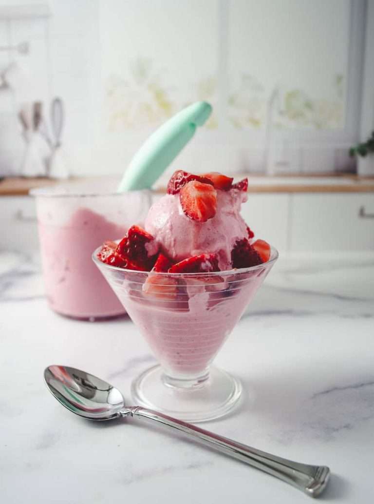 Ninja Ceami Protein Strawberry Ice Cream with Fairlife Milk in a decorative serving cup with cut up fresh strawberries on top, on a white counter with a spoon in front and in the background the pint of ice cream.