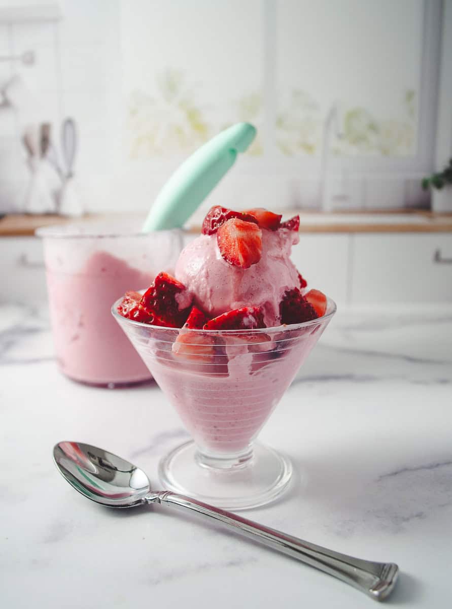 Ninja Ceami Protein Strawberry Ice Cream with Fairlife Milk in a decorative serving cup with cut up fresh strawberries on top, on a white counter with a spoon in front and in the background the pint of ice cream.