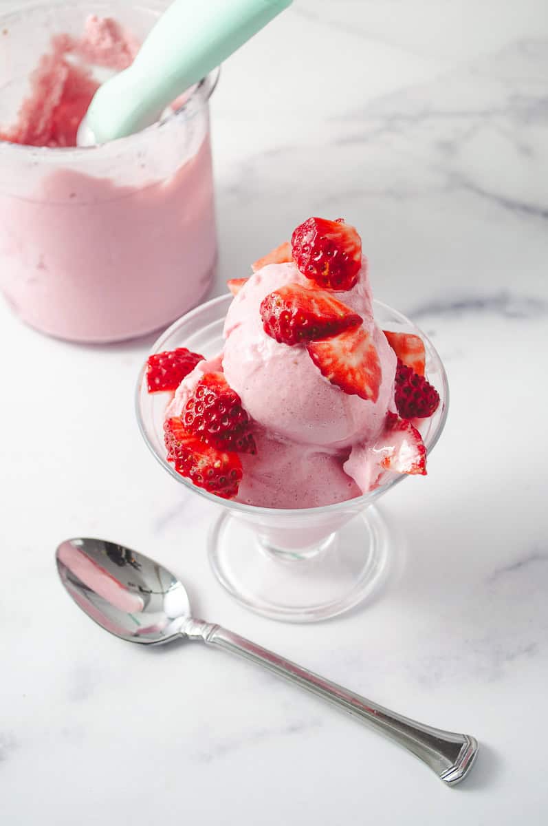 Ninja Ceami Protein Strawberry Ice Cream with Fairlife Milk in a decorative serving cup with cut up fresh strawberries on top, on a white counter with a spoon in front and in the background the pint of ice cream with aqua ice cream scoop in the pint and a top down view. 
