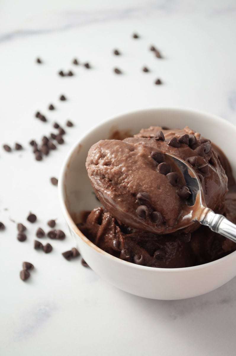 Ninja Creami Protein Chocolate Ice Cream 3 scoop of creamy of ice cream with mini chocolate chips on top, in a white bowl with chocolate chips scattered on the counter, spoon with one a heaping serving of ice cream ready to eat. 