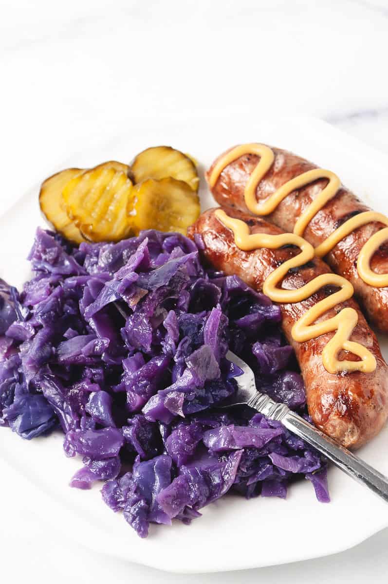 Instant Pot German Red Cabbage with Apple, on w white plate with 2 sausages with honey mustard on top a fork in the cabbage and 5 pickles on the plate.