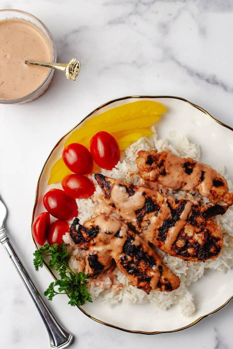 A plate of Bang Bang Chicken over white rice, drizzled with Bang Bang Sauce made with Sriracha Sauce. Garnished with cherry tomatoes, parsley, and yellow bell pepper slices, it’s served on a white plate with a gold rim. A small glass bowl of Spicy Sauce and a fork are nearby on a marble countertop.