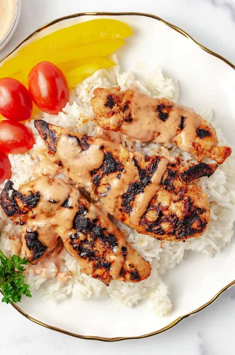 close up of Grilled Bang Bang Chicken on a plate of Crispy Bang Bang Chicken served over white rice, drizzled with Bang Bang Sauce made with Sriracha Sauce. The dish is garnished with cherry tomatoes, parsley, and a side of Spicy Sauce in a small glass bowl, all presented on a white plate with a gold rim against a marble background.