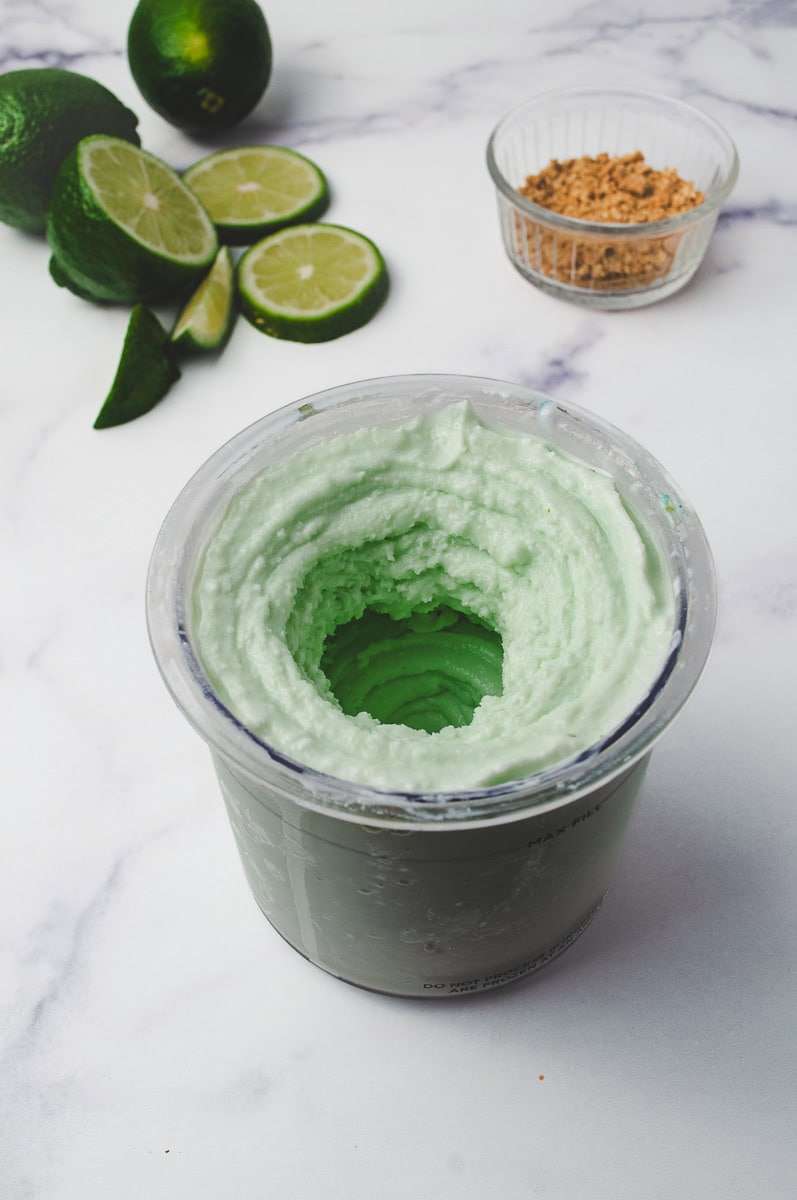 Ninja Creami Protein Key Lime Pie Yogurt Ice Cream in a pint container on a white counter with a lime and lime slices and a small clear bowl of crushed graham crackers. 