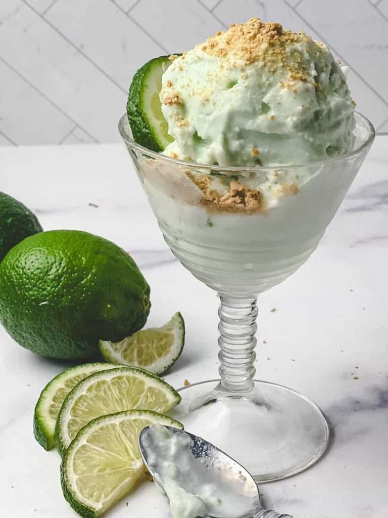 Ninja Creami Protein Key Lime Pie Yogurt Ice Cream 2 scoops in a tall dessert glass graham cracker crumbs on top with s spoon on the marble counter with a splash of ice cream in the spoon a whole lime and 4 slices of lime on the counter with the spoon.