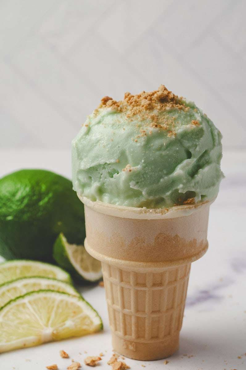 Ninja Creami Protein Key Lime Pie Yogurt Ice Cream in a wafer cone with a whole lime and 4 lime slices on the counter.