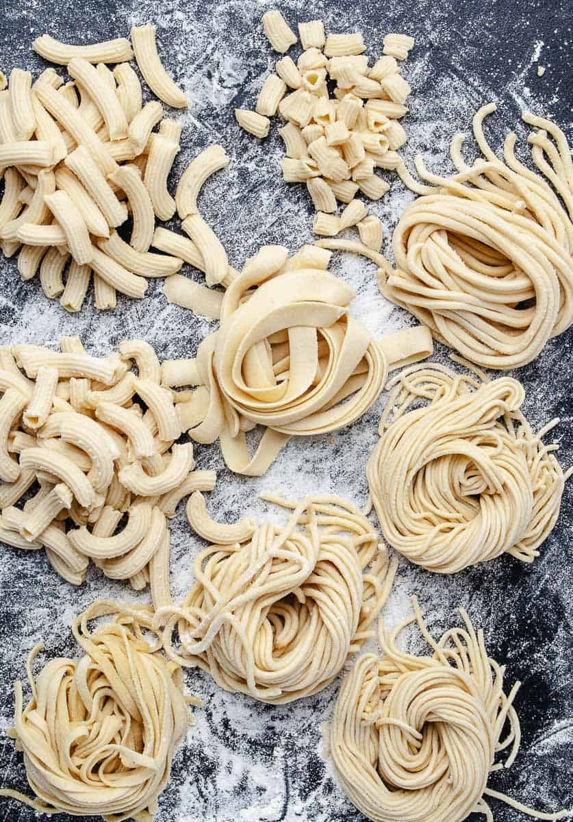 How to Make Homemade Fresh Pasta Using Pasta Maker fresh made 4 swirls of pasta and 2 elbow pasta on a black cutting board with lots of flour to keep them from sticking top view.