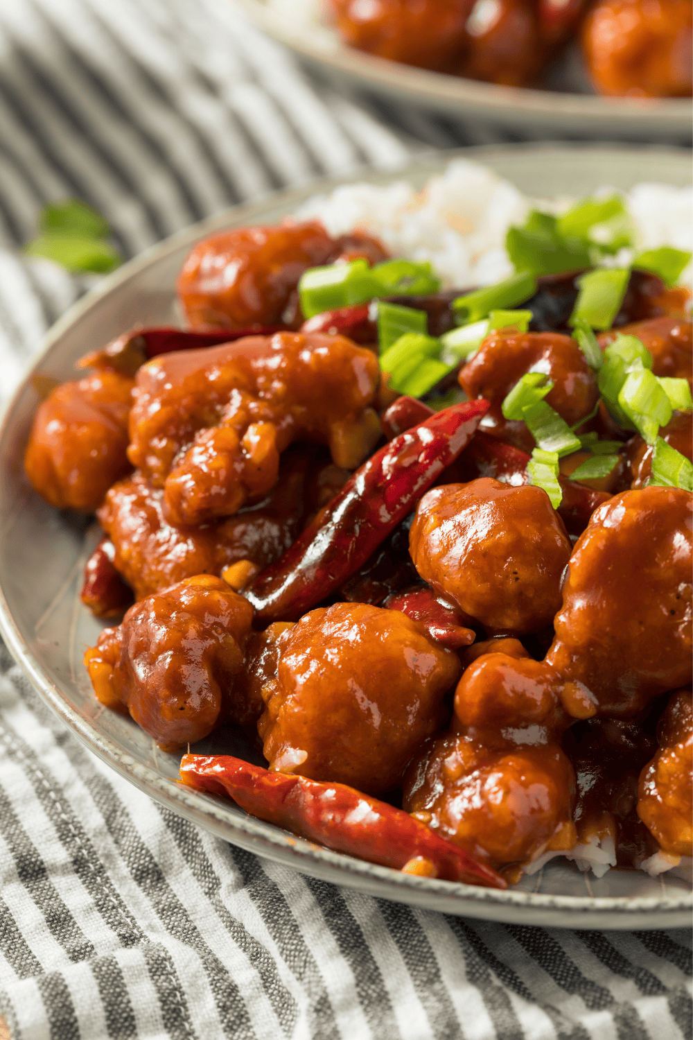 Close-up of General Tso’s Chicken served on a plate with white rice, coated in a glossy spicy sauce, garnished with chopped green onions and whole dried red chilies, placed on a striped cloth background.