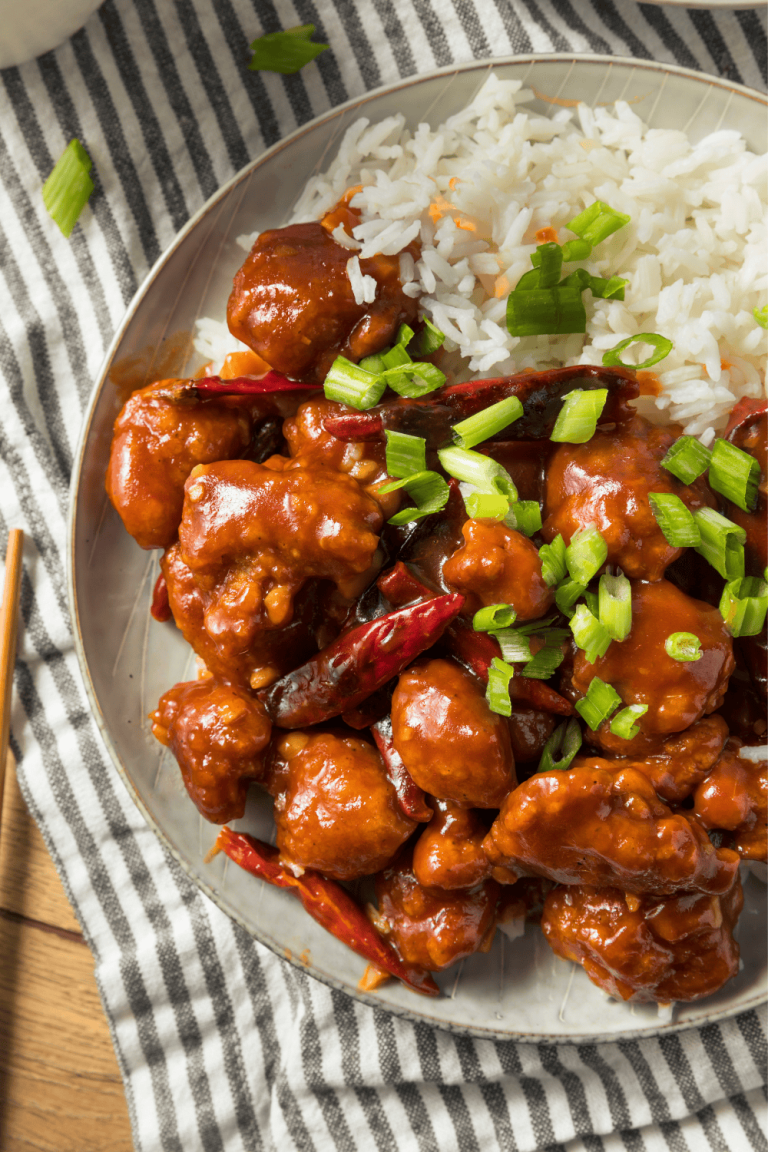 Overhead view of General Tso’s Chicken served with white rice on a gray plate. The chicken is coated in a glossy, spicy sauce and topped with chopped green onions and dried red chilies, placed on a striped cloth.
