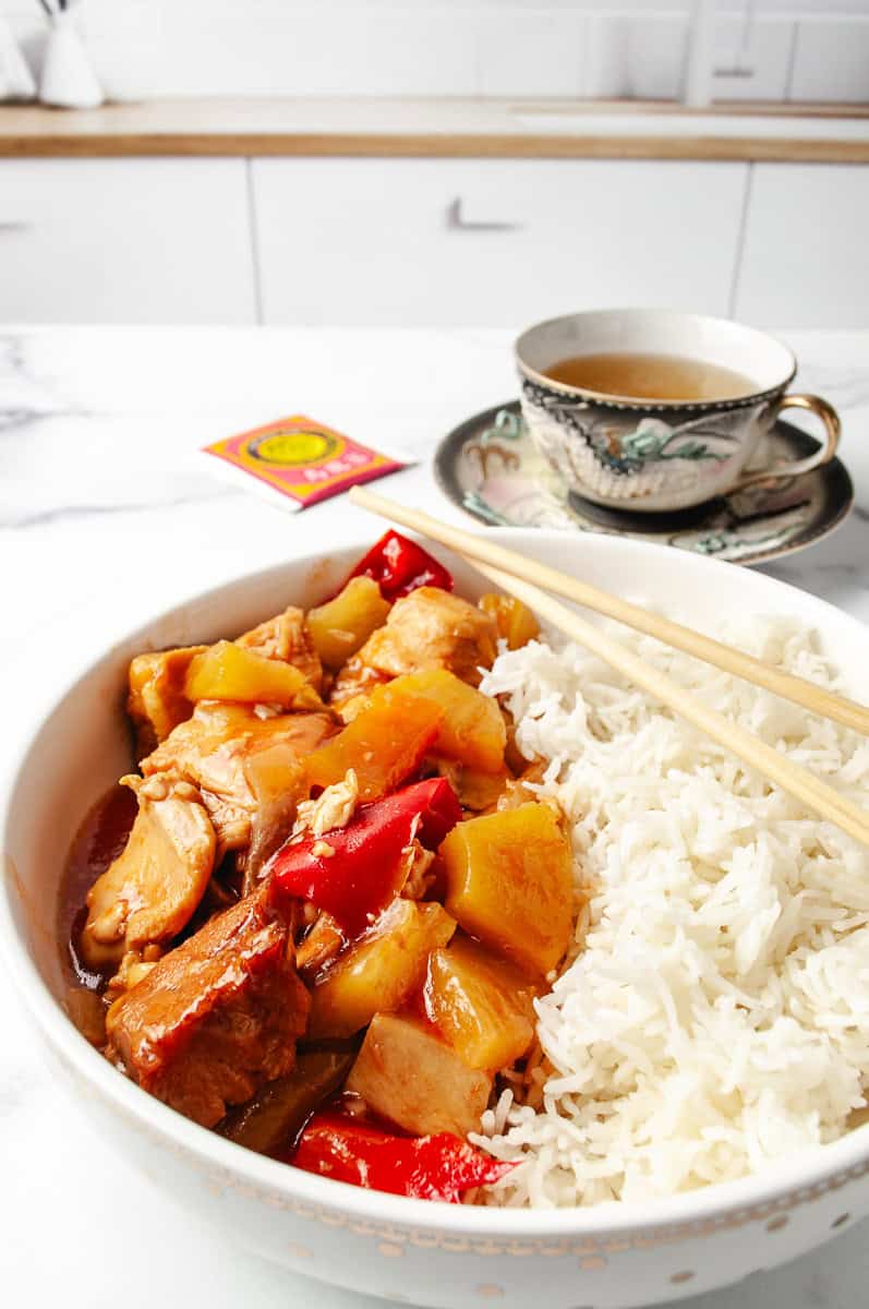 Crockpot Sweet And Sour Chicken served in a bowl with white rice and chopsticks, featuring juicy pineapple, red bell peppers, and tender chicken in a tangy sauce—perfect for Sweet And Sour Chicken Easy, Asian Slow Cooker Recipes, Sweet N Sour Chicken, and other Crockpot Asian Recipes, with a cup of tea and Chinese tea packet in the background.