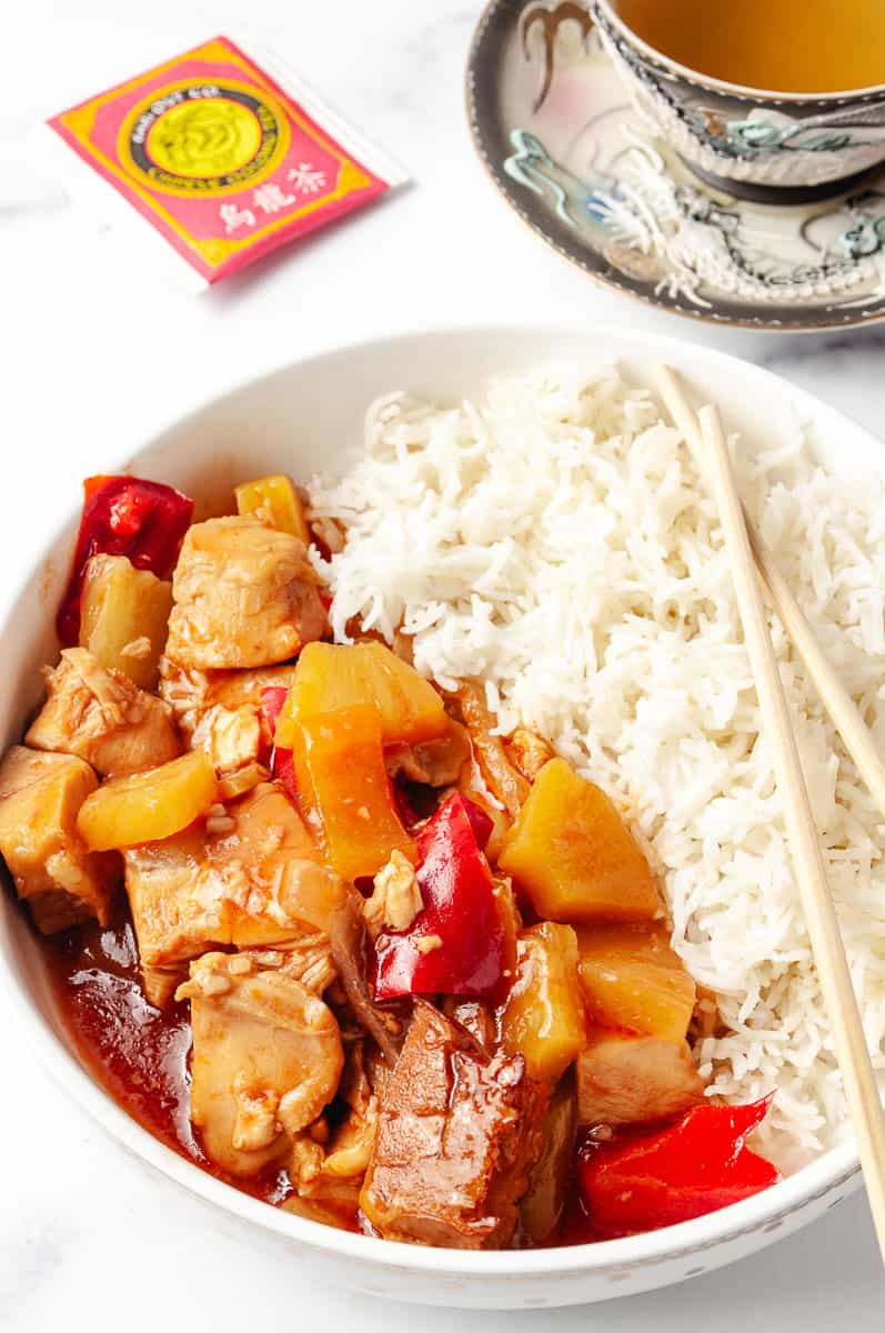 A bowl of CrockPot sweet and sour chicken with pineapple, red bell peppers, and white rice, served with chopsticks. A packet of Chinese oolong tea and a cup of tea in a decorative dragon-themed teacup are beside the bowl.