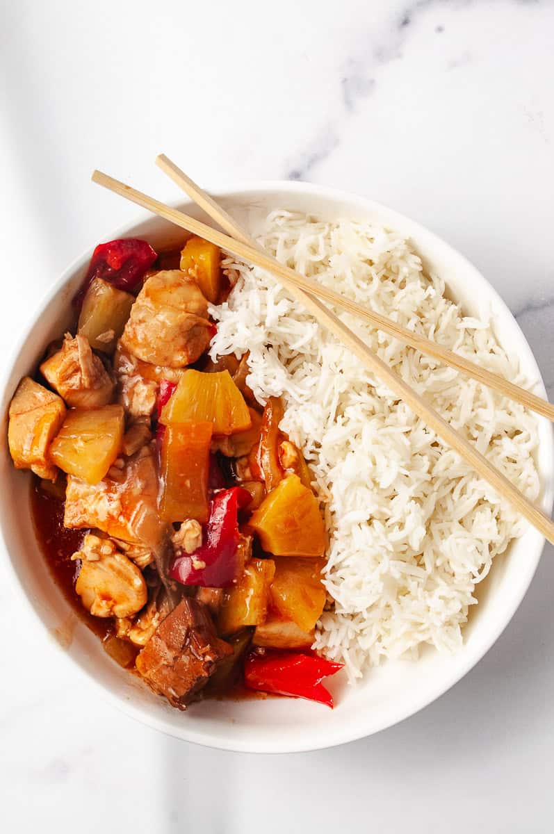 Overhead view of a bowl filled with Crockpot Sweet And Sour Chicken and white rice, featuring pineapple, red bell peppers, and chicken in a glossy sour sauce—ideal for Sweet And Sour Chicken Easy, Crockpot Asian Recipes, Asian Slow Cooker Recipes, and other Sweet N Sour Chicken dishes.