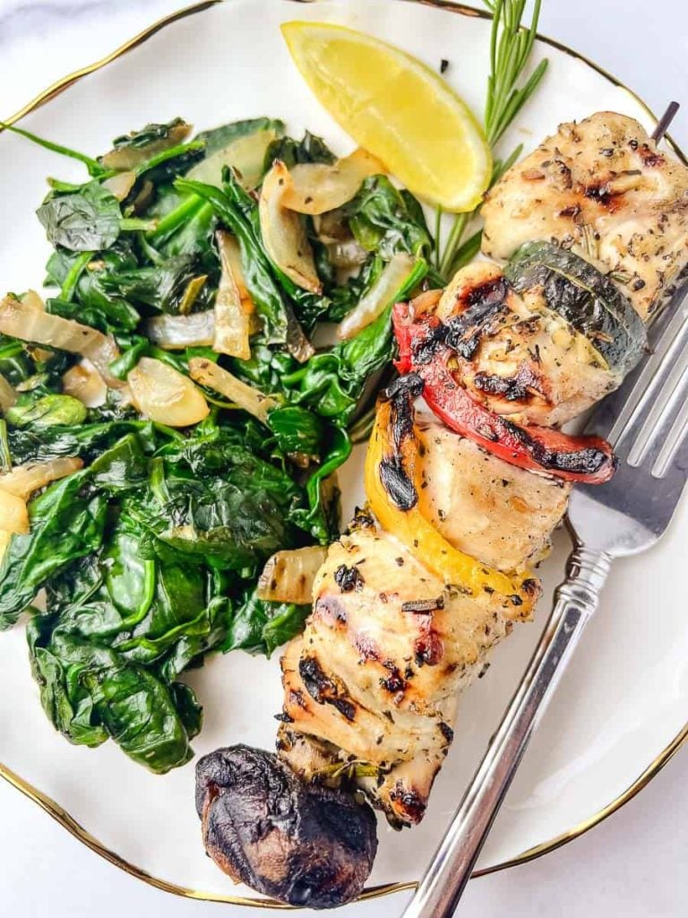 A beautifully plated meal features grilled chicken skewers on the grill, showcasing juicy chicken skewers with colorful bell peppers and mushrooms, served on a metal skewer. The dish is accompanied by a side of sautéed spinach and onions, a quick weeknight meals option. A lemon wedge and rosemary garnish add a fresh flavor, hinting at a delicious chicken skewers marinade easy to prepare.