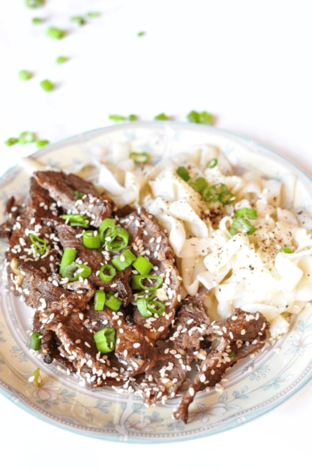 Korean Marinated Sesame Beef Bulgogi on a white plate with flowered rim with sesame seeds on top the green onion with noodles.