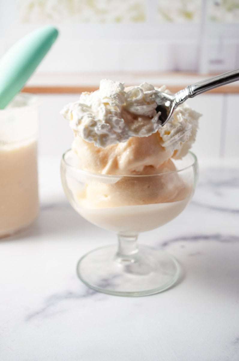 a glass dish filled with scoops of Peach Frozen Yogurt, topped with a generous dollop of whipped cream being scooped out with a spoon. In the background, there's a container of the same Peach Frozen Yogurt with a mint-green scoop resting in it, set against a marble countertop. This delicious treat is made using Ninja Creamy, following a Ninja Ice Cream Recipe, specifically a Homemade Frozen Yogurt and Frozen Yogurt Recipe, highlighting Healthy Peach Recipes. The backdrop features a soft, blurred kitchen setting with light streaming through a window, creating a cozy and inviting atmosphere.