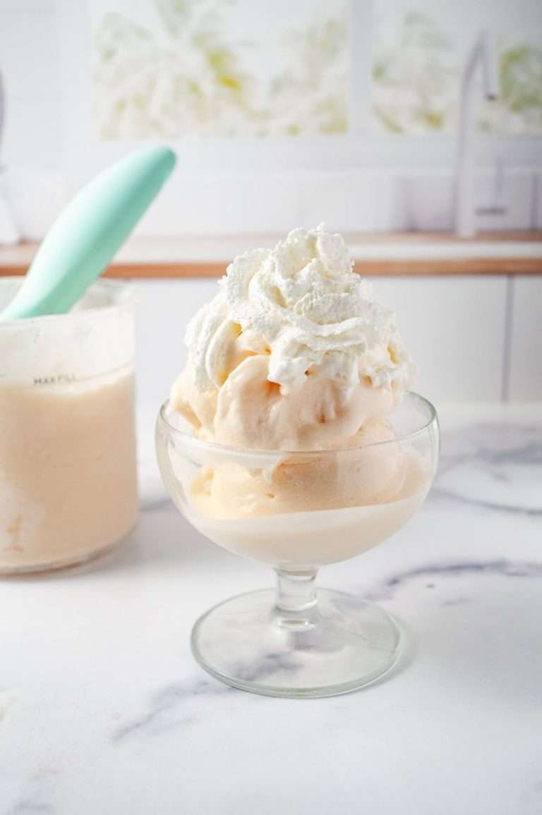 Ninja Creami Peach and Cream Frozen Yogurt Low Sugar a glass dish filled with scoops of creamy peach frozen yogurt, topped with a generous dollop of whipped cream. In the background, there's a container of the same peach frozen yogurt with a mint-green scoop resting in it, set against a marble countertop. The backdrop features a soft, blurred kitchen setting with light streaming through a window, creating a cozy and inviting atmosphere. The dessert looks fresh and delicious, ideal for a refreshing treat, made using a ninja creami recipe for homemade frozen yogurt.