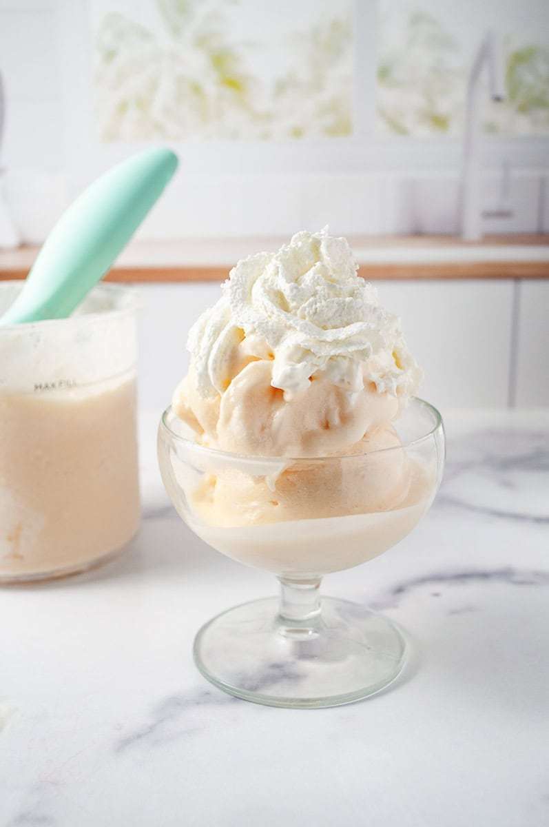 Ninja Creami Peach and Cream Frozen Yogurt Low Sugar a glass dish filled with scoops of creamy peach frozen yogurt, topped with a generous dollop of whipped cream. In the background, there's a container of the same peach frozen yogurt with a mint-green scoop resting in it, set against a marble countertop. The backdrop features a soft, blurred kitchen setting with light streaming through a window, creating a cozy and inviting atmosphere. The dessert looks fresh and delicious, ideal for a refreshing treat, made using a ninja creami recipe for homemade frozen yogurt.