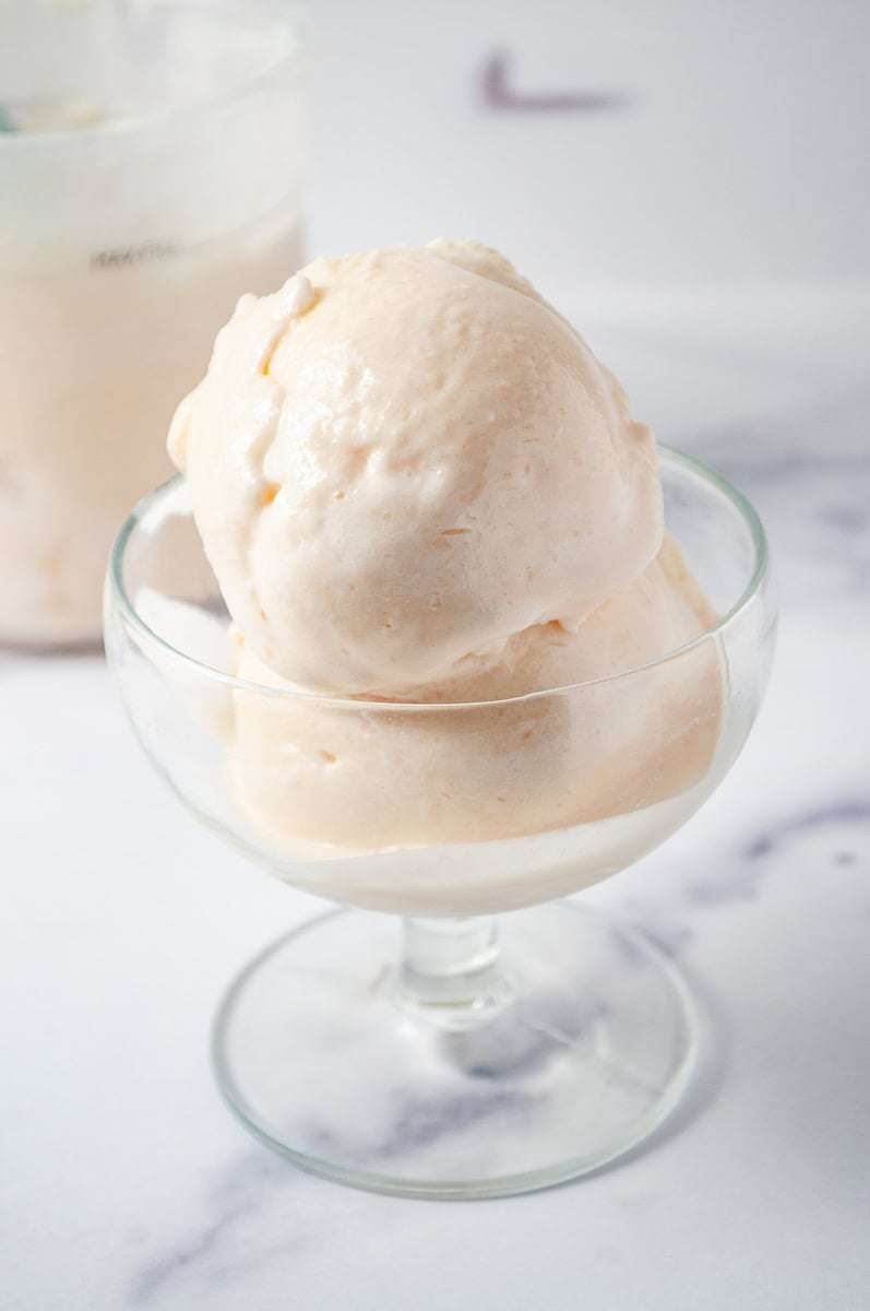 Ninja Creami Peach and Cream Frozen Yogurt Low Sugar a glass dish filled with scoops of Ninja Creami Peach and Cream Frozen Yogurt, a low-sugar homemade treat, topped with a generous dollop of whipped cream. In the background, there's a container of the same peach frozen yogurt with a mint-green scoop resting in it, set against a marble countertop. The backdrop features a soft, blurred kitchen setting with light streaming through a window, creating a cozy and inviting atmosphere. The dessert looks fresh and delicious, perfect for a refreshing, guilt-free indulgence.