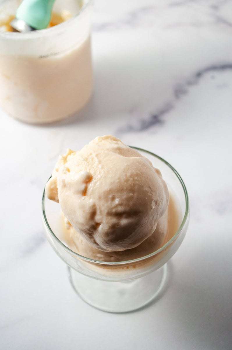 The image shows a top-down view of a glass dish filled with scoops of Peach Frozen Yogurt, a no-sugar-added treat. In the background, there's a container of the same Peach Frozen Yogurt with a mint-green scoop resting in it, set against a marble countertop. This healthy dessert is made using Ninja Creamy, following a Ninja Ice Cream Recipe, specifically a Homemade Frozen Yogurt and Frozen Yogurt Recipe, showcasing Healthy Peach Recipes. The backdrop features a soft, blurred kitchen setting, creating a cozy and inviting atmosphere.