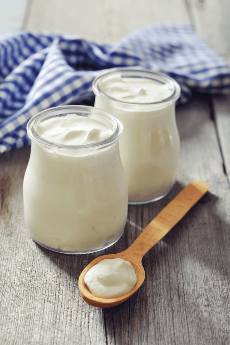 Two glass jars of creamy yogurt on a rustic wooden table with a spoonful in front, perfect for making a tangy and tender Greek yogurt chicken marinade. This simple base works beautifully for yogurt chicken recipes, especially when creating flavorful Greek chicken yogurt dishes or a protein-rich Greek yogurt marinade.
