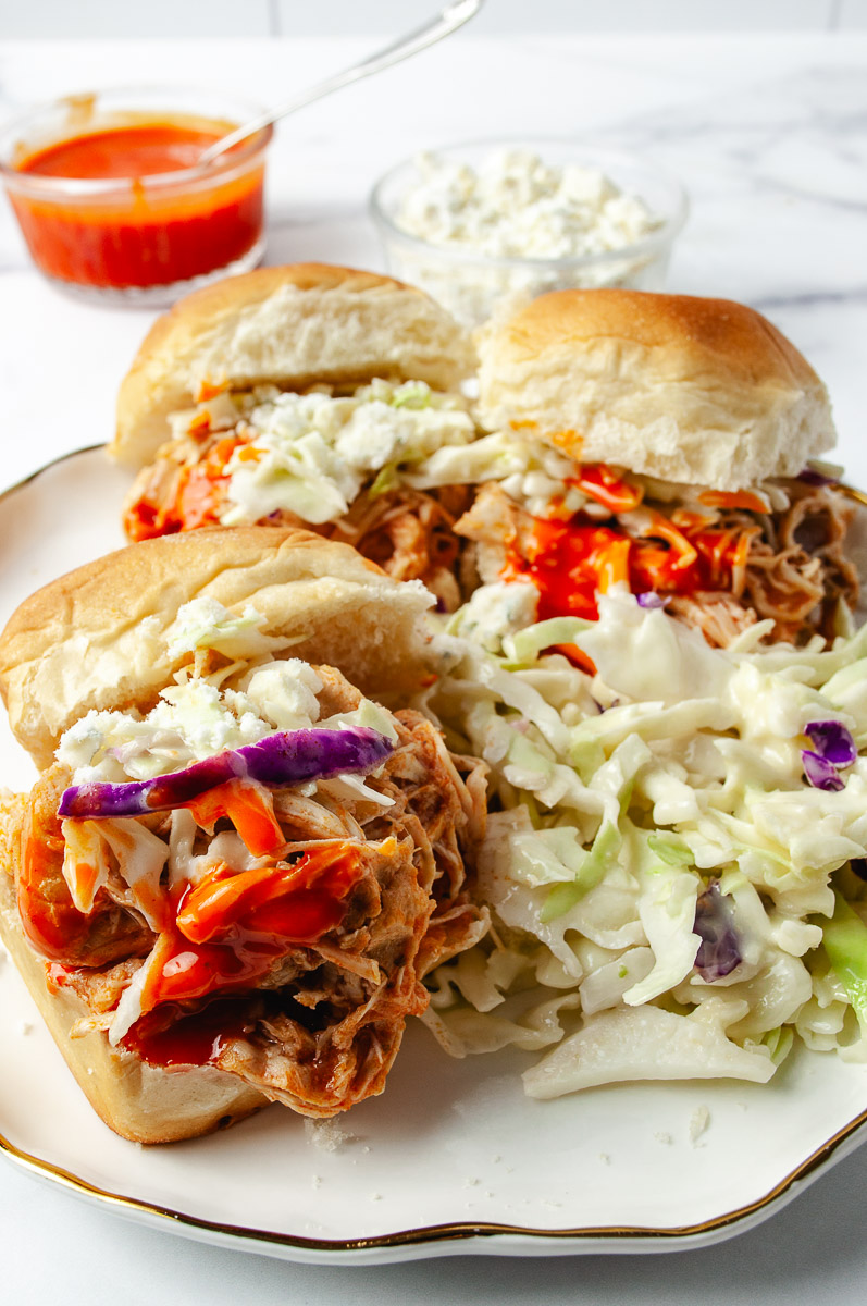 Three Crockpot Buffalo Chicken Sandwiches are stacked on a plate, loaded with shredded chicken tossed in spicy buffalo sauce, topped with creamy slaw, crumbles of cheese, and colorful shredded veggies. A side of crunchy celery sticks chopped into the slaw adds a fresh bite. The sandwiches look like mini versions of buffalo chicken dip on a bun—an easy recipe perfect for the whole family with bold flavor and satisfying texture. In the background, there's a bowl of favorite toppings including extra buffalo sauce and cheese, making this dish ideal for game day or busy weeknights.