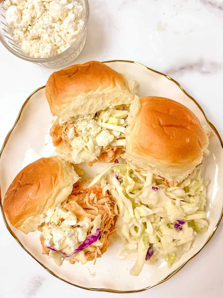 Indulge in the zesty delight of Buffalo Chicken Sliders straight from a Crockpot Buffalo Chicken recipe! These Chicken Sliders feature tender Shredded Buffalo Chicken layered on soft buns, topped with creamy blue cheese crumbles and a refreshing coleslaw mix. A small bowl of extra blue cheese waits on the side for added indulgence.