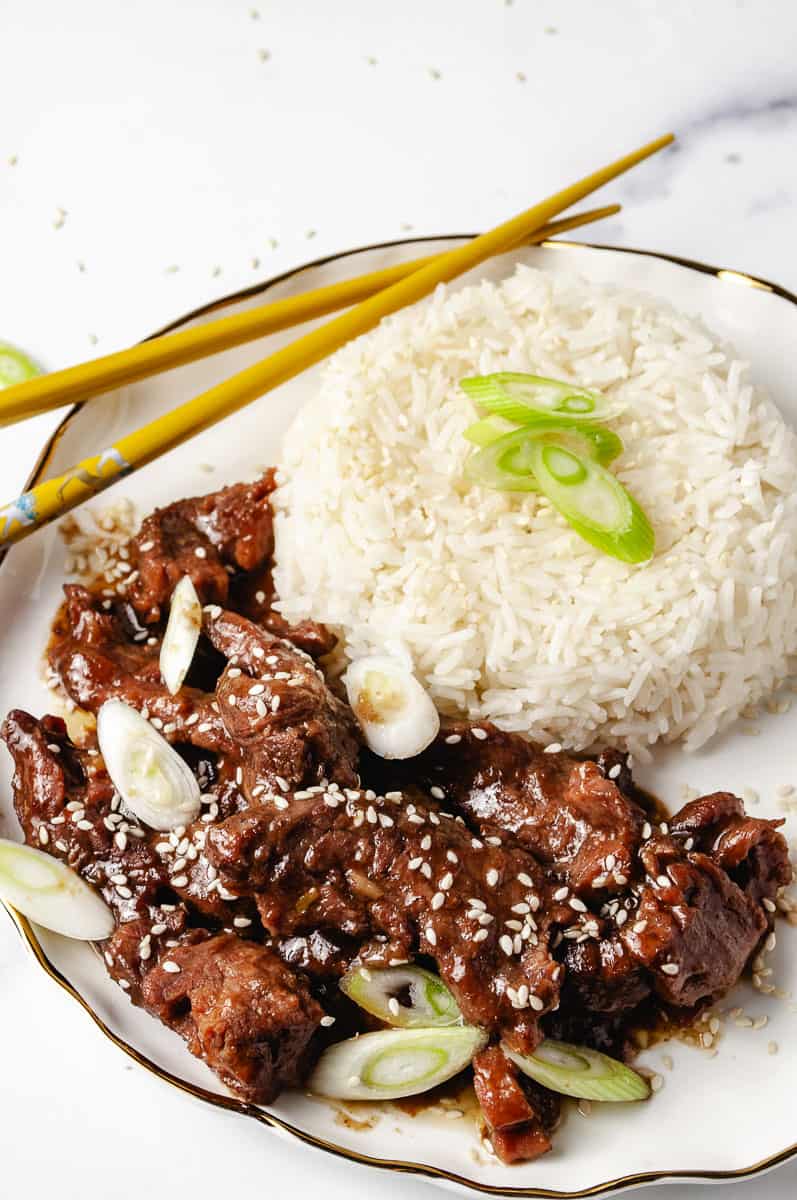 A vibrant plate features tender Mongolian Beef Recipe slow-cooked in a Crock Pot Slow Cooker, served alongside a mound of fluffy white rice topped with sliced green onions and sesame seeds. The rich, glossy sauce coats the Beef Dishes, with yellow chopsticks adding a pop of color, making this Crockpot Mongolian Beef Recipe a hearty family meal.