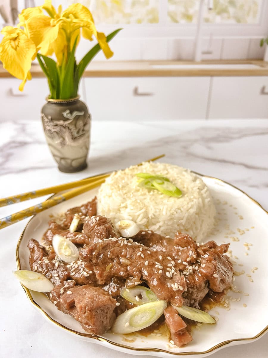 A delightful plate of Mongolian Beef Crock Pot features tender beef pieces in a rich, glossy sauce, garnished with sesame seeds and green onions. It’s served alongside a fluffy mound of white rice topped with fresh green onion slices, presented on a white plate with a gold rim. Yellow chopsticks and a vase of bright yellow irises add a cheerful touch on a marble kitchen countertop, making this Beef Mongolian Recipe a cozy meal.