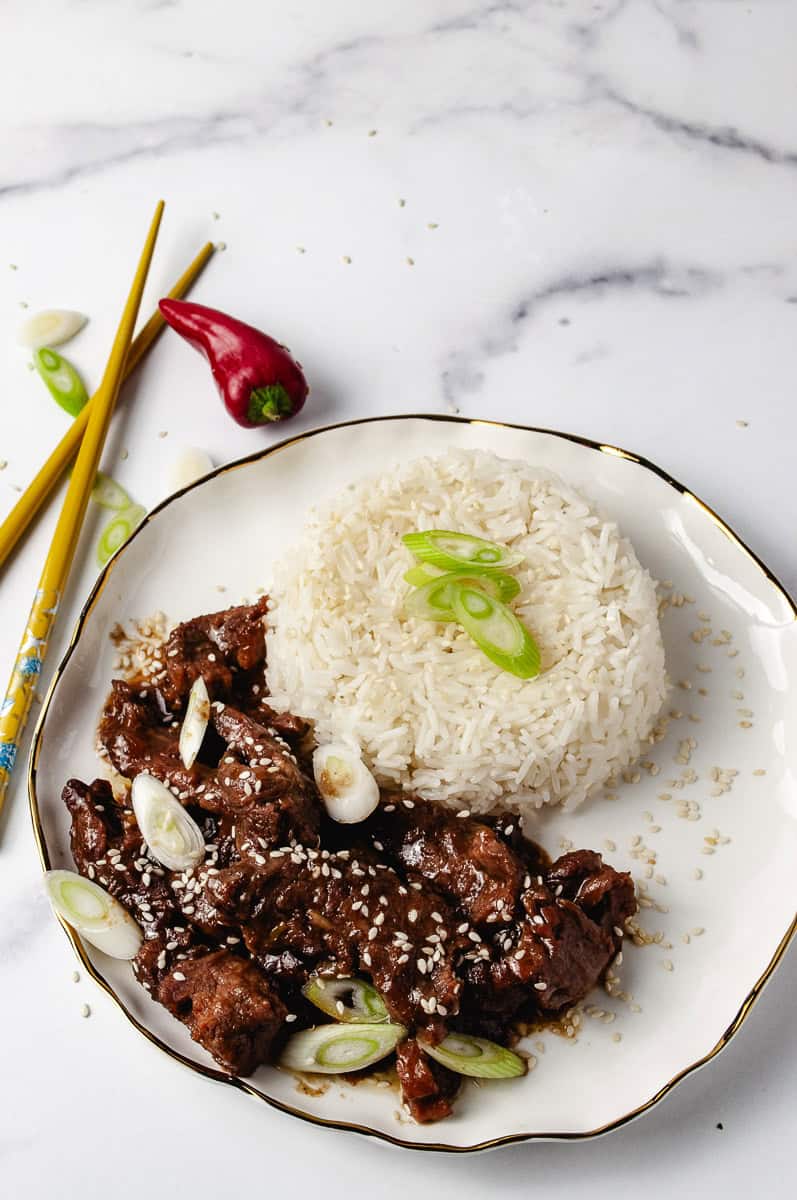 A stunning plate of Mongolian Beef Slow Cooker features succulent beef chunks in a glossy, savory sauce, sprinkled with sesame seeds and green onions. Served next to a mound of fluffy white rice topped with fresh green onion slices, this Crock Pot Mongolian Beef Recipe is accented with yellow chopsticks and a vibrant red chili pepper on a white marble surface, highlighting the Beef Dishes’ bold flavors.