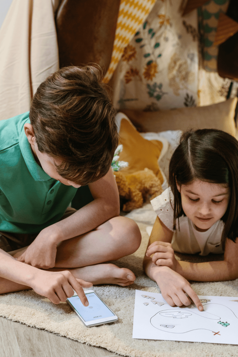 Two kids sitting on the floor playing a printable summer scavenger hunt game together. One child points at a scavenger hunt list on paper, while the other uses a phone to follow along. Perfect example of fun indoor or outdoor scavenger hunt for kids.