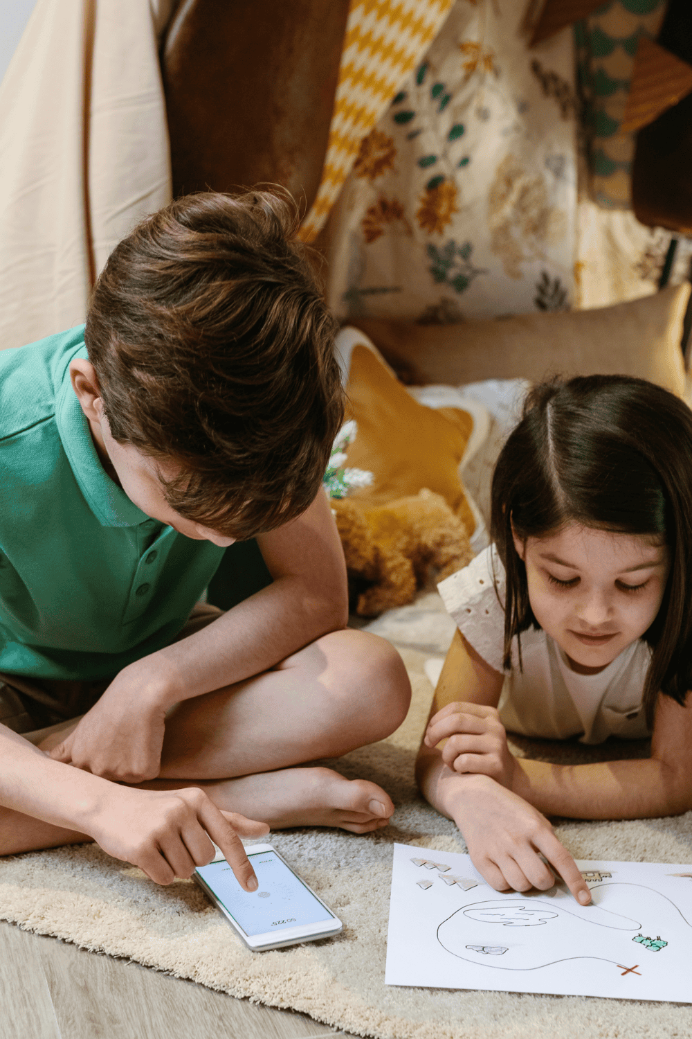 Two kids sitting on the floor playing a printable summer scavenger hunt game together. One child points at a scavenger hunt list on paper, while the other uses a phone to follow along. Perfect example of fun indoor or outdoor scavenger hunt for kids.