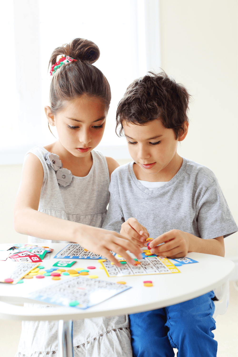 Two young children play a colorful bingo game at a small white table, using markers and cards. This printable game for kids is perfect for screen free activities at home or on a road trip.