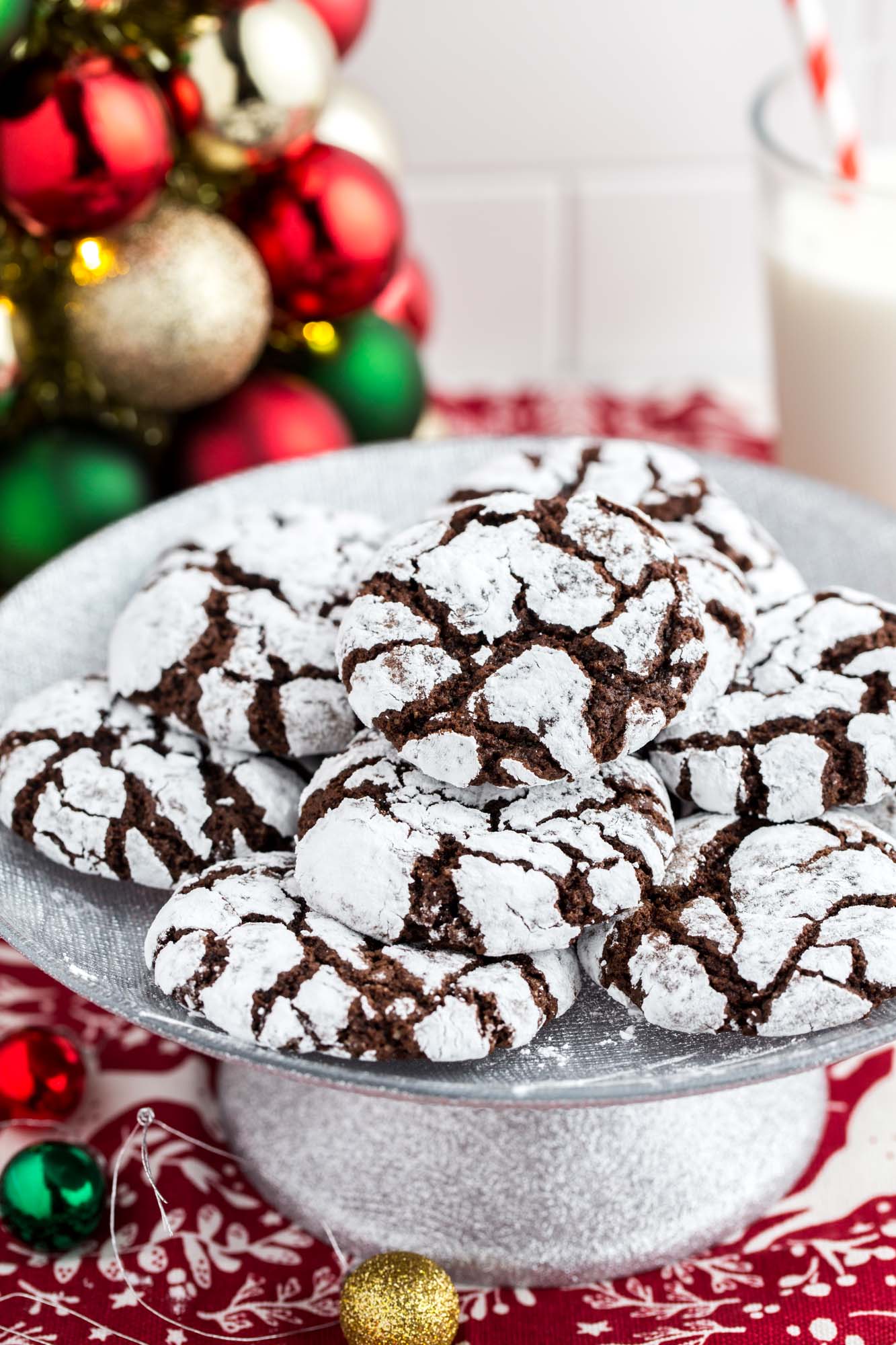 A festive plate of chocolate crinkle cookies coated in powdered sugar, stacked on a silver cake stand. The cookies have a rich dark chocolate color with crackled white tops. Christmas ornaments in red, green, and gold decorate the background, along with a glass of milk and red striped straw, creating a cheerful holiday cookie scene.