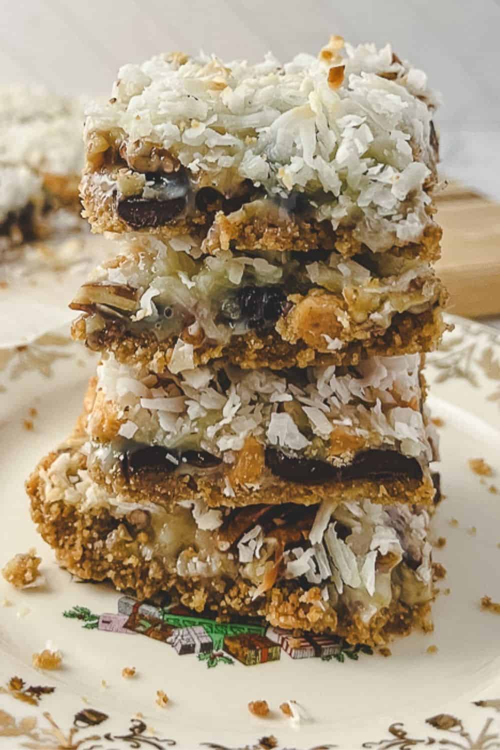 Easy 7 Layer Magic Cookie Bars stack of 5 squares on a gold flowered plate the 10 other cookie bars on parchment on a wood cutting board.