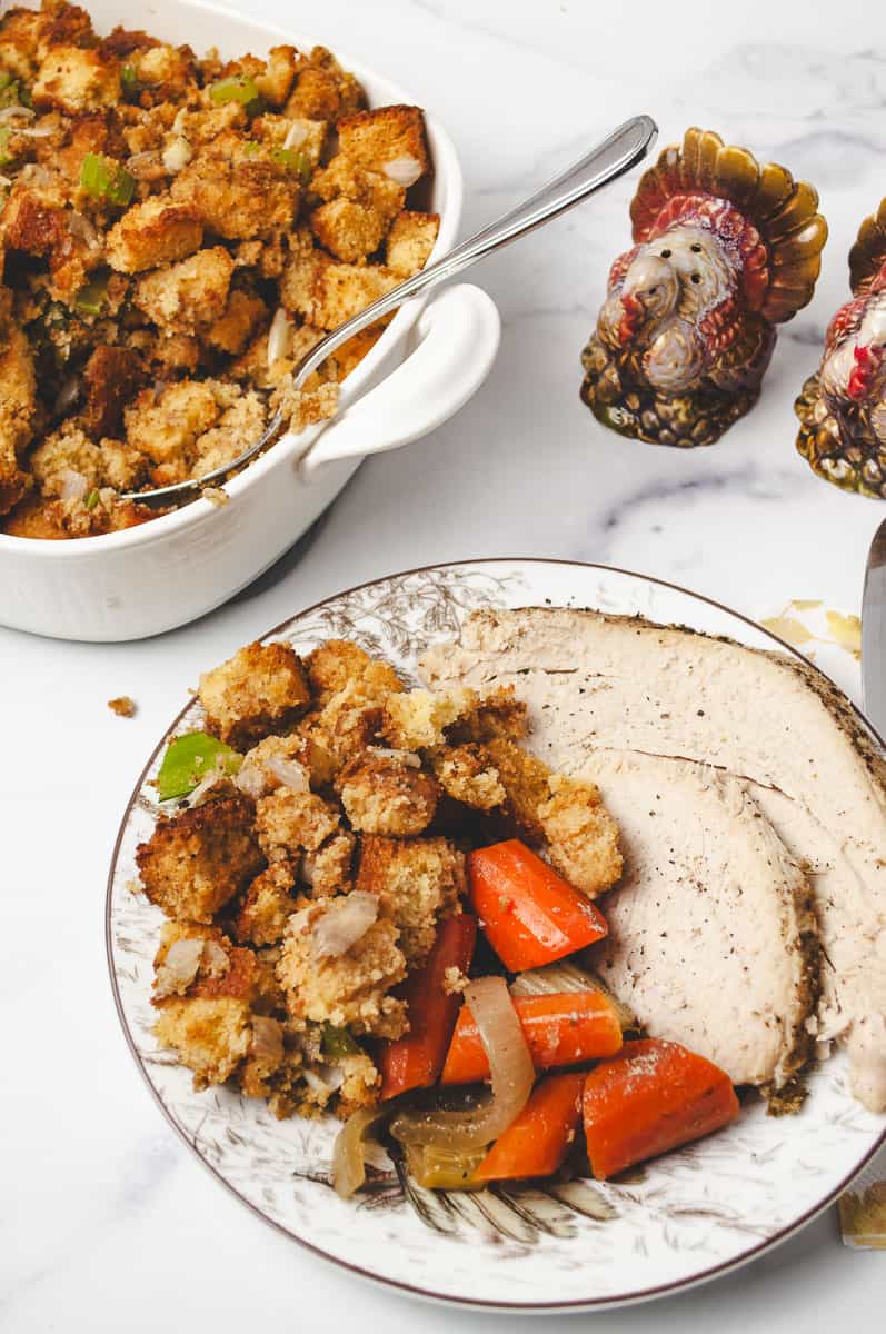 Plate of roasted turkey, glazed carrots, and Liberty Tree Tavern-style cornbread stuffing with celery and onion. Served beside a white dish of homemade cornbread dressing and Thanksgiving turkey salt and pepper shakers. A cozy meal perfect for cornbread stuffing recipes and holiday stuffing recipes.