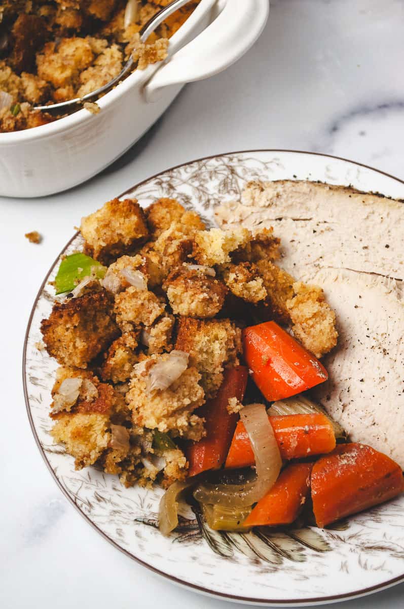 Plate of homemade cornbread stuffing with tender roast turkey and cooked carrots, served next to a white dish of cornbread dressing. A cozy holiday meal featuring Liberty Tree Tavern-style cornbread stuffing, perfect for stuffing recipes and cornbread easy dinner ideas.