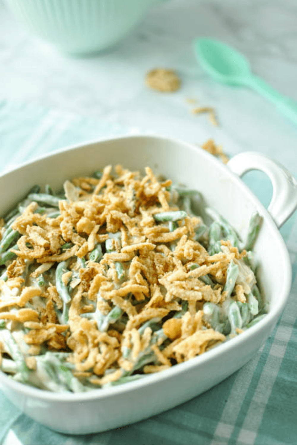Green bean casserole topped with crispy fried onions in a white baking dish, sitting on a teal plaid cloth with a mint green spoon in the background.