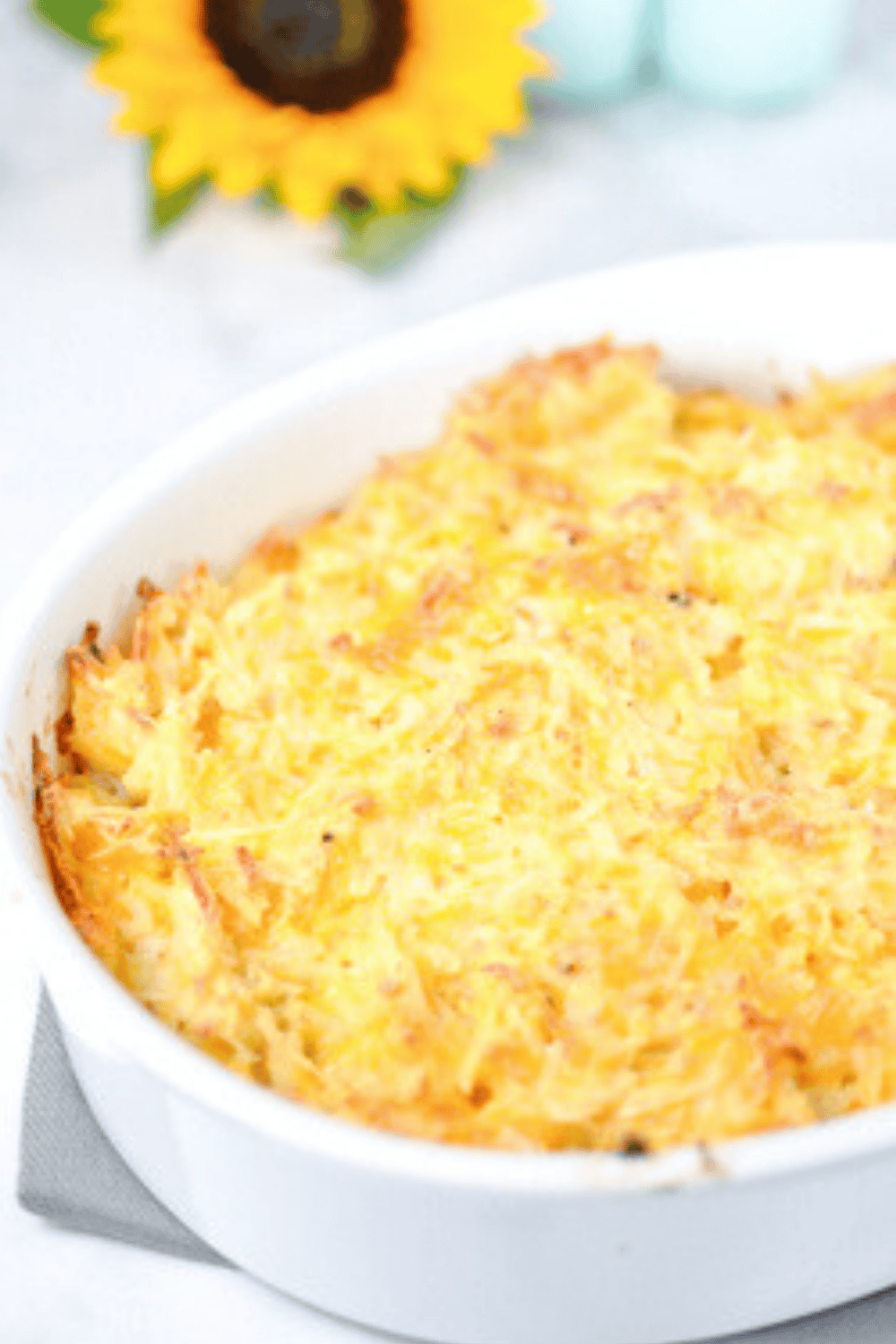 Golden-brown cheesy hash brown potato casserole baked in a white dish, with a bright sunflower and soft-focus background decor.